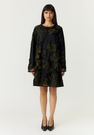 Femme debout portant une robe noire mi-longue avec une broderie florale verte et des chaussures à talons noires sur un fond uni.