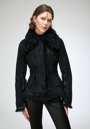 Femme aux cheveux courts et bruns porte une veste noire cintrée avec un grand col duveteux et un pantalon noir assorti sur un fond uni.