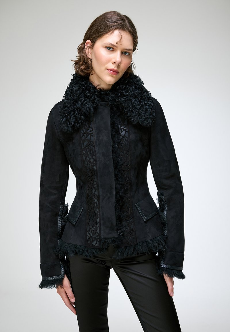 Femme aux cheveux courts et bruns porte une veste noire cintrée avec un grand col duveteux et un pantalon noir assorti sur un fond uni.