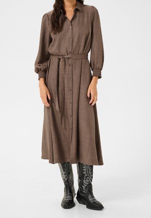 Kobieta w brązowej, zapinanej na guziki sukience midi z paskiem w talii i czarnych kowbojskich butach z haftem, stojąca na jednolitym tle.