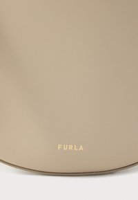 Μπεζ δερμάτινη τσάντα με λεία υφή, διακοσμημένη με χρυσό λογότυπο "FURLA" στο μπροστινό μέρος. Απλή, στρογγυλεμένη μορφή χωρίς επιπλέον μεταλλικά στοιχεία.