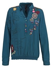 Blouse à manches longues bleu sarcelle avec broderie florale multicolore, lettres en patchwork sur la poitrine, boutons colorés et ceinture élastique.