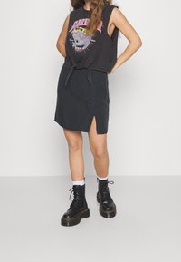 Černý bezrukávový top s potiskem a uzlem v přední části, kombinovaný s černou sukní ke kolenům s bočním rozparkem a černými šněrovacími botami.