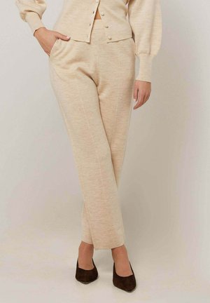 Vrouw die beige gebreide broek en bijpassend vest met pofmouwen draagt, staand met één hand in de zak en donkerbruine puntige schoenen.