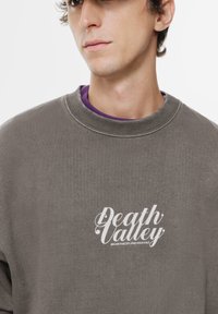 Szürke pulóver, kerek nyakkal, fehér "Death Valley" felirattal és egy szlogennel. Puha tapintású és laza fazonú, lila pólót visel alatta.