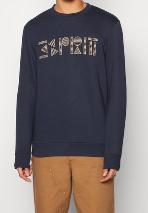Granatowa bluza z napisem "Esprit" w geometryczny wzór, noszona z beżowymi spodniami przez stojącą osobę na jednolitym tle.