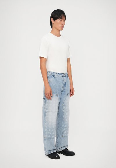 Camiseta blanca y pantalones de mezclilla azul claro de gran tamaño con patrones sutiles y múltiples bolsillos, combinados con zapatos negros.