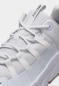 Λευκό αθλητικό παπούτσι Nike Free με πλεκτό επάνω μέρος, μοναδικό σύστημα δέσιμου κορδονιών και χοντρή σόλα, δείχνοντας κοντινές λεπτομέρειες υφής και σήμανσης.