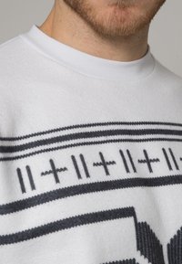 Bílá mikina s měkkou texturou, zdobená černým geometrickým vzorem a pruhy přes hrudník a ramena. Kruhový výstřih.