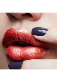 Metallisch oranje-rode lippen met een opvallende textuur en glans, die vollheid uitstralen. Een donkere paarse nagel raakt de lippen aan, wat de kleurcontrasten benadrukt.