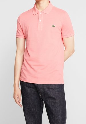 Mann trägt ein hellrosa kurzärmeliges Poloshirt mit kleinem grünem Krokodil-Logo auf der Brust und dunkelblaue Jeans.