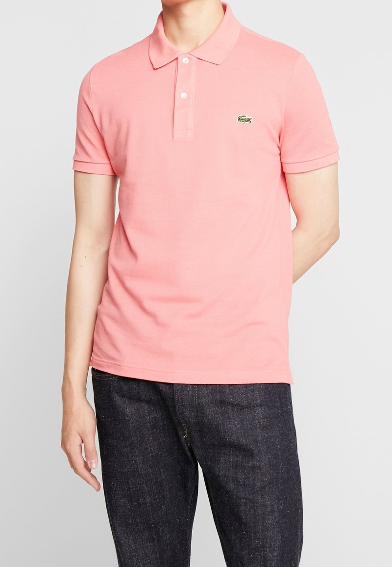 Homme portant un polo à manches courtes rose clair avec un petit logo crocodile vert sur la poitrine et un jean bleu foncé.