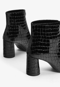 Botas negras, brillantes hasta el tobillo con una textura de cocodrilo, que cuentan con una puntera redondeada y tacones cilíndricos gruesos. Sin herrajes visibles.