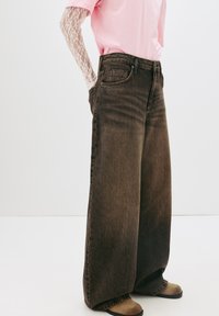 Rjave široke jeans hlače z visokim pasom, dizajnom s petimi žepi in rahlimi madeži. Ustrezajo z rožnato srajco in čipkastimi dolgimi rokavi.