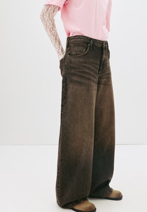 Jean large marron à taille haute, design à cinq poches, avec un léger délavage. Associé à une chemise rose avec des accents en dentelle à manches longues.