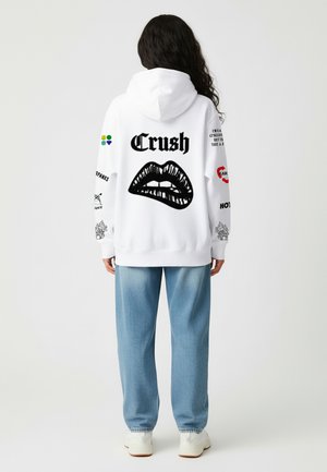Weißer Kapuzenpullover mit einem schwarzen Lippen-Graphic und dem Text "Crush" auf dem Rücken, sowie verschiedenen Druckdetails an den Ärmeln.