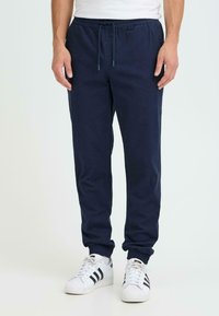 Pantalon de jogging marine avec poignets et taille élastiques. Tissu en coton lisse avec fermeture à cordon. Associé à des baskets blanches avec des rayures noires.