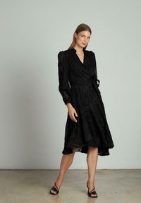 Schwarzes Wickelkleid mit langen Ärmeln, strukturiertem Blumenmuster, ausgestellt Saum und gebundenem Taillenband. Getragen mit schwarzen Riemenabsätzen auf einem beigen Boden.