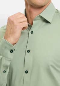 Hellgrünes Hemd aus glattem Stoff, mit dunklen grünen Knöpfen und einem spitzen Kragen. Die Manschetten haben Knopfdetails.
