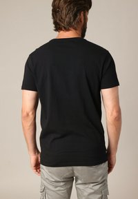 Homme aux cheveux bruns et à la barbe, portant un t-shirt noir simple à manches courtes et un pantalon cargo gris clair, de dos sur un fond neutre.