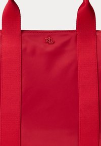 Raudona tote dėžė iš audinio, su dviem plačiais rankenų ir įspaustu logotipo detaliu, glotnia tekstūra, siuvinėjimo akcentais ir paprastu dizainu.
