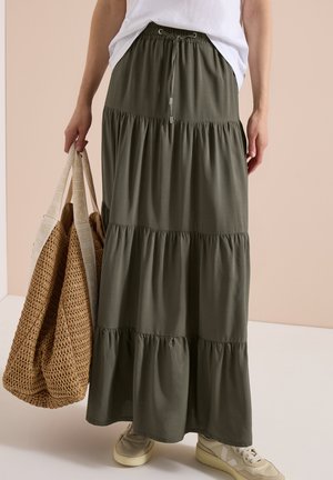 Kobieta w długiej oliwkowozielonej rozkloszowanej spódnicy, białych tenisówkach, trzymająca dużą plecioną torbę typu tote z materiałowymi uchwytami.