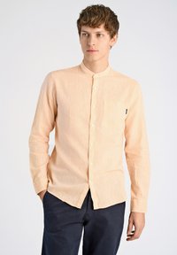 Licht perzikkleurig overhemd met een mandarin kraag, een enkele borstzak en een subtiele textuur. Gecombineerd met donkerblauwe broek.