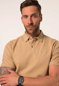 Béžová polokošile s krátkým rukávem, má límeček a jemný emblém jelena. Model má na zápěstí černé hodinky s texturovaným páskem.