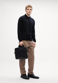 Μαύρο πουλόβερ με κουμπιά, καφέ παντελόνι, μαύρα βραδινά παπούτσια. Μοντέλο κρατά μια μαύρη δερμάτινη τσάντα με διπλές λαβές και μπροστινή τσέπη, μινιμαλιστικός σχεδιασμός.