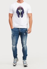 Camiseta blanca de manga corta con un gráfico de un faraón con rayas moradas, combinada con jeans azules desgastados y zapatillas grises.