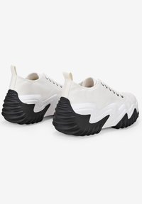Sneakers bianchi in tela con suola spessa nera e ondulata, caratterizzati da un design con lacci, linguetta e punta tonda. Accenti laterali testurizzati.