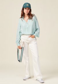 Camicetta a righe verde menta con bottoni, pantaloni bianchi a gamba larga, cappellino verde con logo e sneaker bianche; tiene una borsetta piccola con una tracolla a righe.