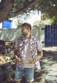 Camicetta a maniche corte, rosa con motivi floreali e blu, abbinata a pantaloni corti in denim chiaro. Ambientazione naturale con un albero e un muro sullo sfondo.