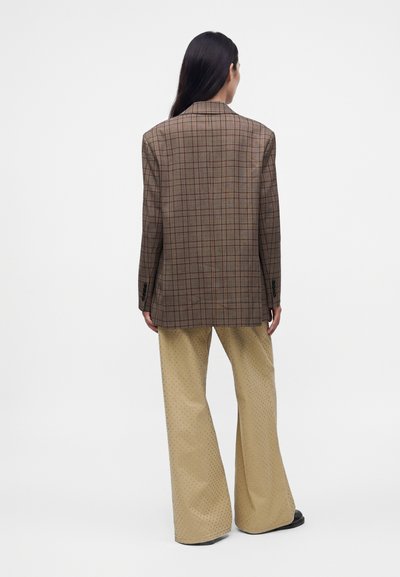 Femme aux longs cheveux foncés portant un blazer marron à carreaux et un pantalon larges beige de dos sur un fond blanc uni.