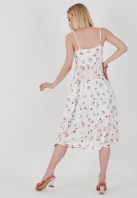 Blumiges Midikleid aus leichtem weißen Stoff. Mit dünnen Trägern, gestuften Design und verstreuten Pastellblumenmustern; kombiniert mit roten Absatzmules.