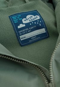 Vihreä fleece-vuorattu takki, jossa on sininen vedenpitävä tarra, jossa on pilvikuva. Sisältää vetoketjun ja pehmeän tekstuurin mukavuutta varten.