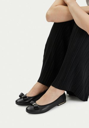 Persona con pantalones negros plisados y bailarinas negras con pequeños lazos, sobre un fondo claro, sentada con los brazos apoyados sobre las rodillas.