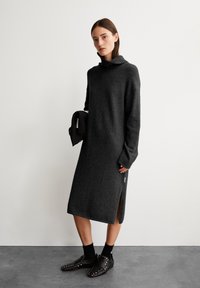 Robe en tricot gris foncé avec un col montant, des manches longues, des fentes sur les côtés et une texture douce, associée à des chaussures à enfiler noires à clous et un sac noir.