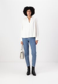 Hvit langermet bluse med V-hals og innsamlet detalj, kombinert med lyseblå jeans og svarte ankelstøvler, holder en lysegrå håndveske.