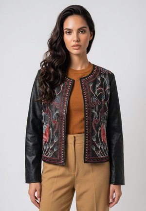 Mujer con cabello largo y oscuro lleva una chaqueta de cuero negra con coloridos patrones bordados de pájaros sobre una blusa marrón y pantalones beige.