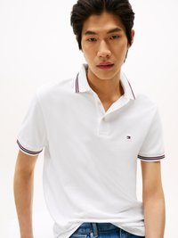 Weißes Polohemd mit einer Knopfleiste mit zwei Knöpfen und kontrastierenden roten und marineblauen Streifen am Kragen und an den Ärmeln. Getragen mit blauen Jeans.