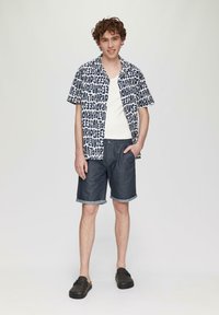 Chemise à manches courtes avec un motif de texte noir et blanc, associée à un short en denim foncé avec des ourlets retroussés, et des chaussures à enfiler sombres.