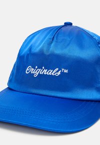 Zila satīna cepure ar izliekto malu, priekšpusē ar baltu izšūtu "Originals™" logotipu. Gluda tekstūra un strukturēts dizains.