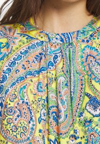 Zbliżenie na szyję kobiety oraz wzorzysty materiał z kolorowymi motywami paisley i kwiatowymi w odcieniach niebieskiego, pomarańczowego, zielonego i żółtego.