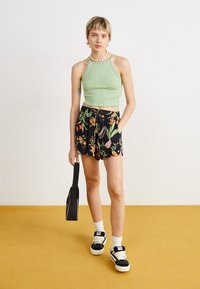 Helle grüne gerippte Crop-Top, schwarze Shorts mit buntem Blumenmuster, schwarze Tasche, weiße Socken, schwarze Sneakers mit weißen Sohlen.