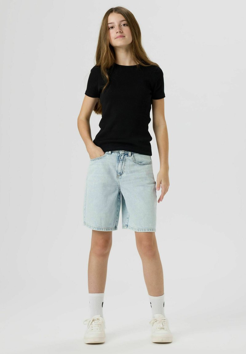 Jeune personne aux cheveux longs portant un t-shirt noir, un short en jean clair, des chaussettes blanches et des baskets blanches, debout avec une main dans la poche.