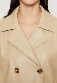 Vrouw in een beige dubbelzijdige leren jas met brede revers en twee grote bruine knopen, zichtbaar van schouders tot bovenste borst.