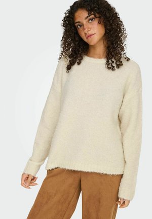 Femme aux cheveux bouclés portant un pull en laine crème et un pantalon marron, se tenant devant un fond clair.