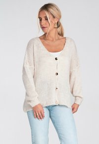 Jasno beżowy dzianinowy kardigan z dekoltem w serek, pięcioma guzikami z przodu i ściągaczami na rękawach. Posiada luźny krój oraz fakturowany wzór.