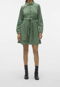 Žena v kolenním zeleném kordovém šatech na knoflíky s opaskem na zavázání a černými kotníkovými botami na podpatku.